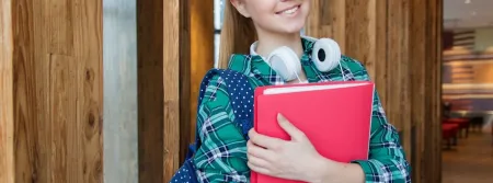 A young woman standing in the hallway, holding school books and smiling at the camera