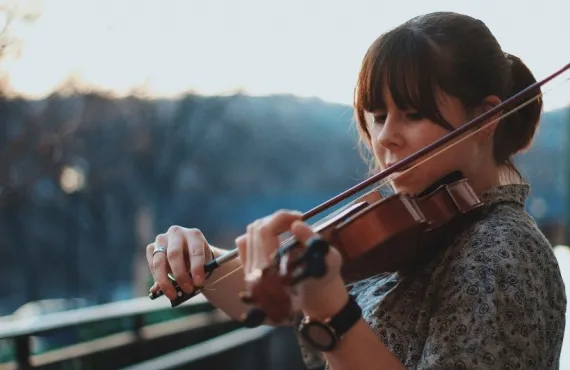 A young woman playing violin on a balcony with trees in the background