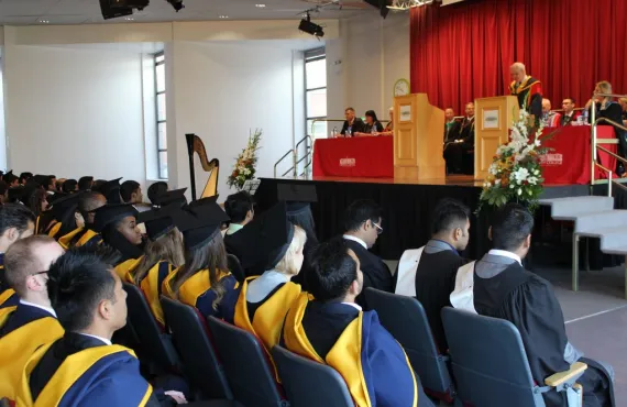Several rows of graduates in cap & gowns look up at a raised dais with a speaker at a Griffith College graduation ceremony