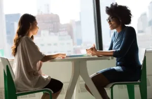Two women sitting at a table in front of a window, conducting an interview