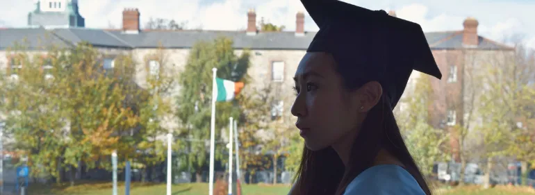 A student in graduation cap and gown standing at a window, with the main Dublin campus in the background.