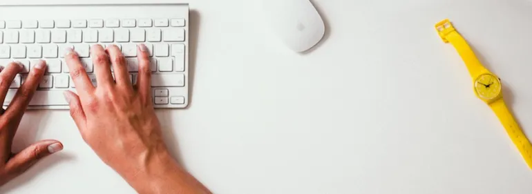 Hands typing on a keyboard on a white desk, with a mouse and a bright yellow watch.
