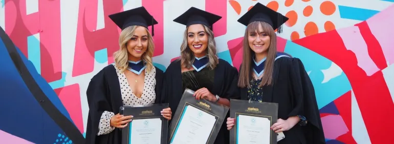Three graduates standing in front of the "happy" mural on Griffith College's main Dublin campus