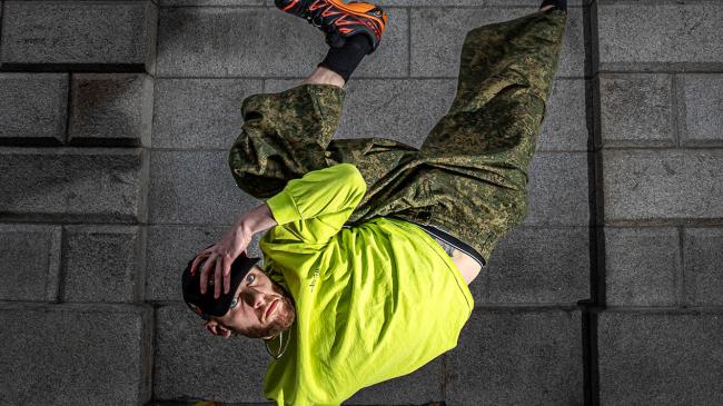 A person in a neon yellow hoodie performs a one-handed freeze, legs kicked up in the air, against a gray stone archway.