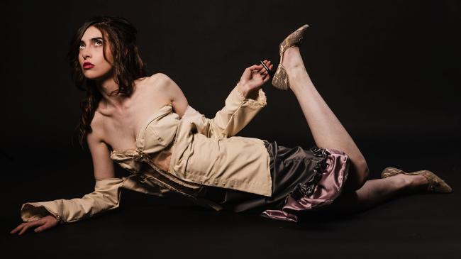 Model lying on the floor, wearing a grey satin skirt and a beige corset with silk and lace details, against a black backdrop