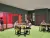 A 3D architectural render of a modern classroom with dark green walls and a light green patterned floor. The room is furnished with circular yellow tables that have integrated navy steel benches. Dark wood doors and a window are visible in the background.