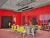 A 3D architectural render of a vibrant classroom featuring bold red walls and a patterned orange floor. Eight small tables connected together with a wooden finish, surrounded by bright yellow chairs. Two sleek black linear pendant lights hang from the ceiling.