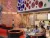 A 3D architectural render of an indoor canteen area. The scene features a red brick wall with a neon flamingo sign and the text CHILL, EAT, DRINK. Circular wooden tables with integrated seating are set on a light green patterned floor, and modular red and navy hexagonal benches are positioned in the foreground.