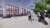 This image shows a 3D architectural visualization of the exterior of The Beacon House, showing a covered bike shelter to encourage sustainable commuting. The render shows the industrial building with large, red-framed windows, people interacting in the exterior, sheltered area for bicycle parking.
