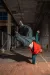 A person in an orange puffer vest and green hoodie performs a one-handed freeze in an urban walkway, legs kicked high toward the ceiling.