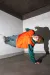 A person in an orange vest and green outfit balances on one hand, body angled sideways, casting a sharp shadow on a white concrete wall.