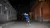 A person in a blue tank top performs a handstand in a graffiti-lined alley at night, brick buildings flanking both sides.