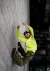 A person in a neon yellow shirt and camo pants mid-jump against a gray stone wall, shot from a tilted angle with city buildings behind.