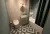 Image shows a hotel guest bathroom render with patterned floor tiles, blue-green walls, art deco wall covering, brass details, vanity unit, toilet, bathtub, shelving and a dark green shower curtain. 