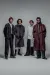 Four young men stand in a minimalist studio against a plain grey background, wearing contemporary, oversized fashion. Their outfits include loose suits, layered shirts, shorts with long socks, and a long maroon coat, all styled in muted tones. They face the camera with neutral expressions, spaced evenly in a slightly staggered formation.