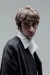 Close-up portrait of a young man with tousled light brown hair, wearing an oversized dark blazer over a white collared shirt. He faces slightly to the side while looking directly at the camera with a neutral, serious expression, set against a plain grey background.