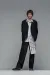 Full-body studio portrait of a young man standing against a plain gray background, wearing an oversized black suit with wide-leg trousers and a white shirt featuring an abstract linear pattern. He faces forward with a neutral expression, hands behind his back, and one leg slightly bent.