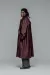  A full-length fashion portrait of a man with dark dreadlocks looking over his shoulder against a solid grey background. He is wearing a voluminous, oversized burgundy trench coat with a storm flap on the back, paired with wide-leg charcoal grey trousers and chunky black leather boots.