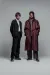A full-length fashion portrait of two men standing side-by-side against a solid grey background. The man on the left wears a structured, collarless black blazer over a white button-up shirt, paired with wide-leg black trousers and chunky black boots. The man on the right wears an oversized, voluminous burgundy trench coat over a grey shirt, charcoal wide-leg trousers, and black leather boots. 
