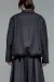 A medium shot from the back of a person with shoulder-length brown hair, wearing a structured, charcoal grey textured jacket and matching voluminous pleated trousers or skirt. The jacket has a gathered, elasticated hem that creates a rounded silhouette, set against a plain grey background.