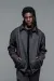  A medium fashion portrait of a man with dark dreadlocks standing against a solid grey background. He is wearing a structured, dark grey textured zip-up jacket with a collar and his hands in his pockets. The jacket is worn over a black shirt and paired with matching grey trousers.