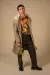 Male model stands in studio wearing a long grey herringbone overcoat with a floral shirt, holding a yellow rose in front of a beige background