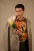 Male model in herringbone overcoat with sheepskin fur collar and floral shirt, holds a yellow rose against a beige background