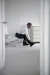 A person in formal clothes sits on a chair fastening a shoe in a bright minimalist room.