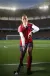 A hockey player in a red uniform crouches on a floodlit pitch, holding a hockey stick with a ball at her feet.