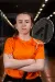A badminton player in an orange shirt stands with folded arms, holding a racket in an indoor sports hall.