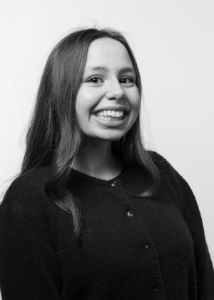 A black-and-white studio portrait of a young woman with long hair, wearing a dark cardigan and smiling warmly at the camera.