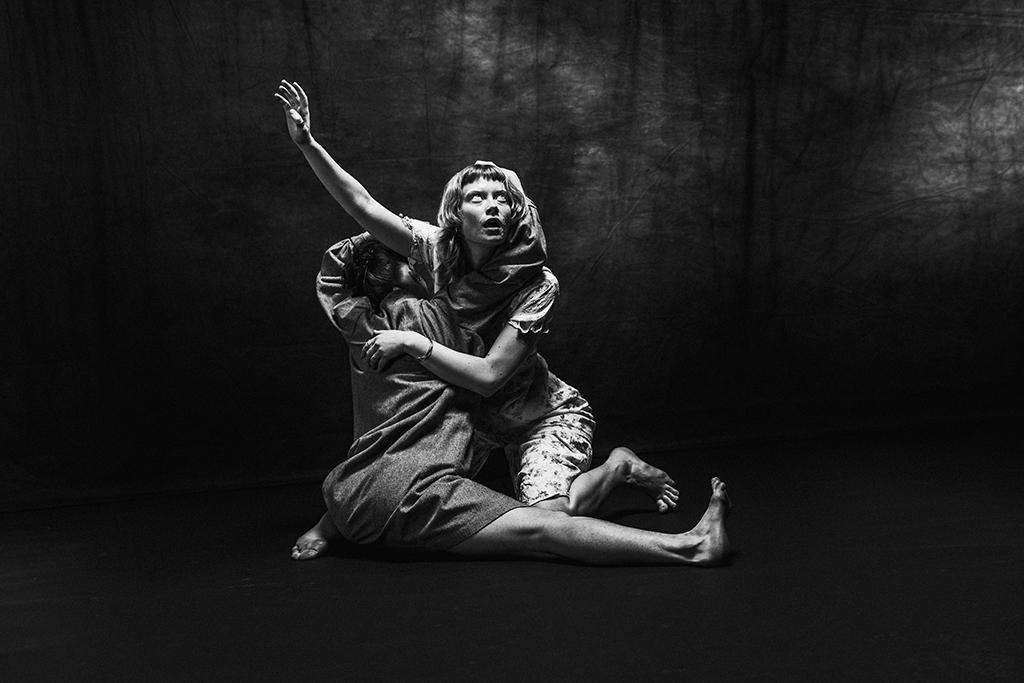 Black and white image of dramatic staged pose with two performers intertwined on a dark background.