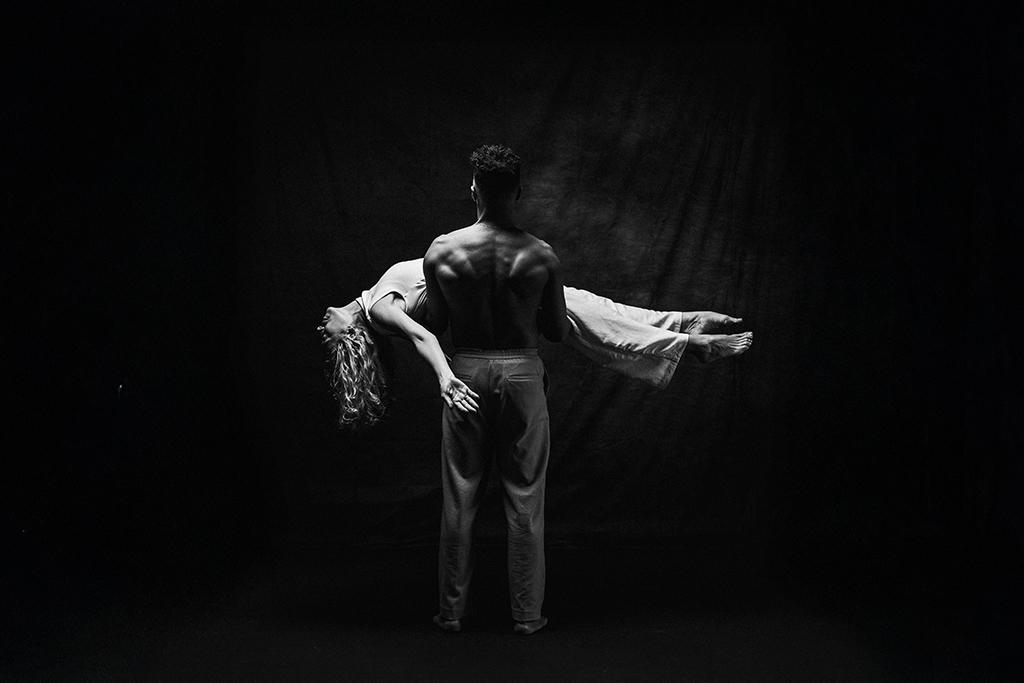 Black and white image of a performer standing and holding another performer horizontally across their arms on a dark stage.
