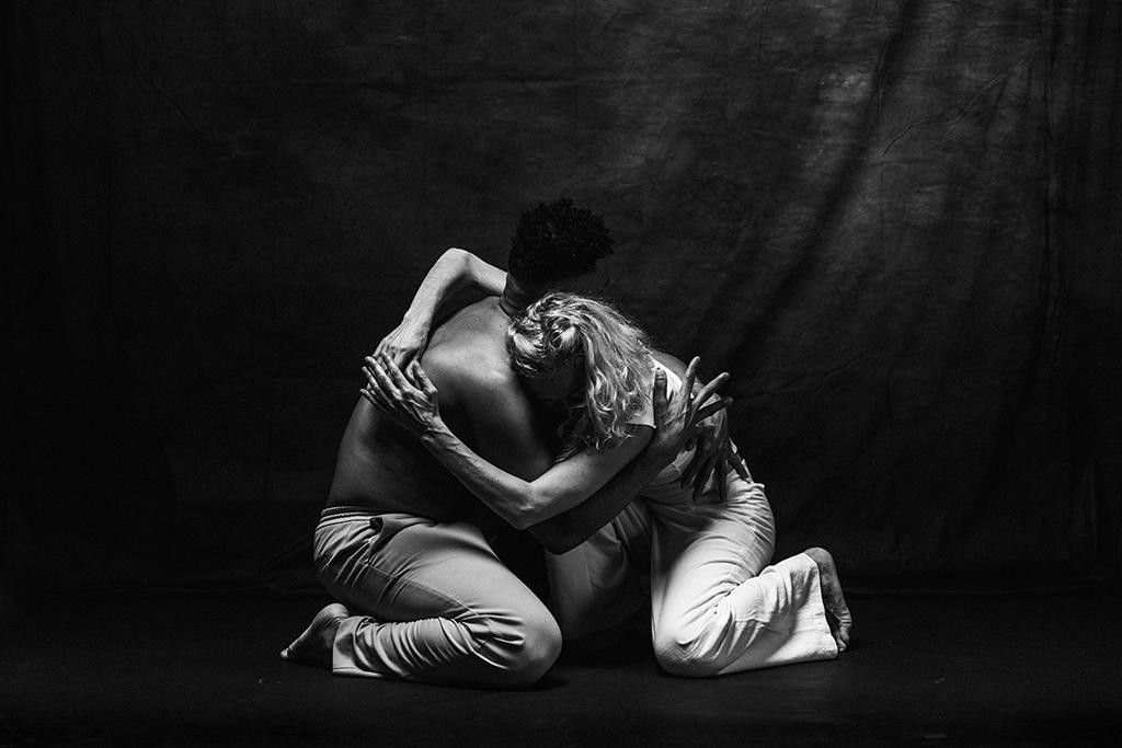  Black and white image of two performers kneeling and embracing tightly on a dark stage.