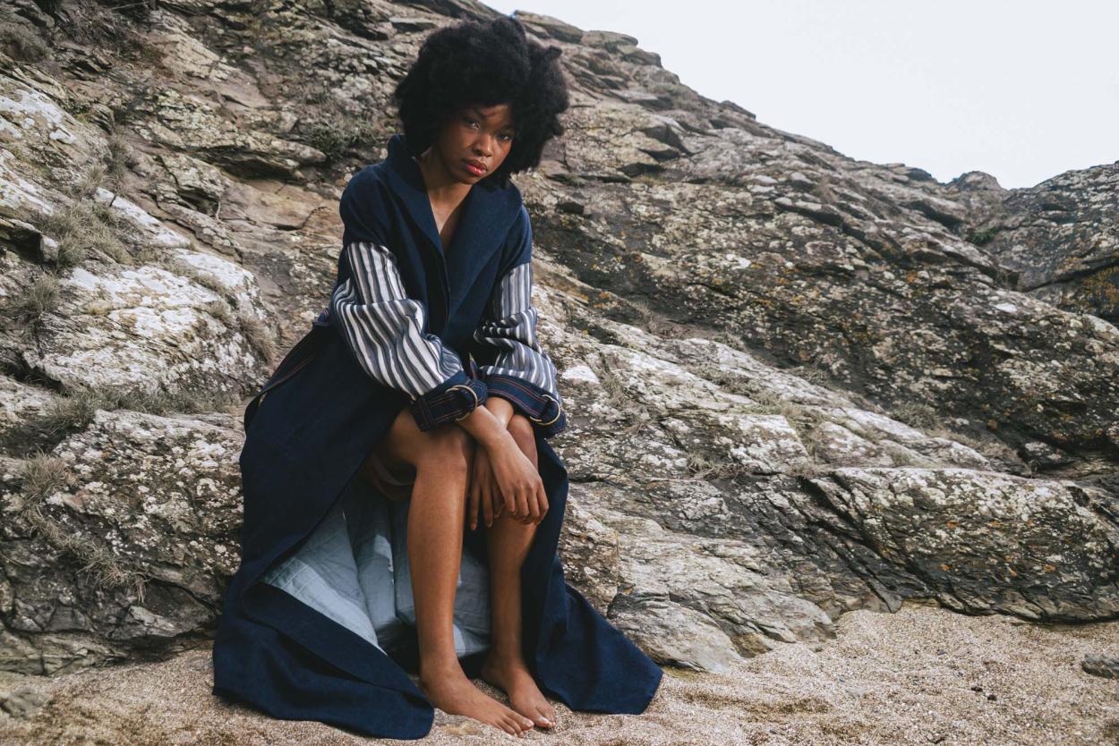 Model sitting on rough coastal rock, in long denim coat with stripe silk chiffon embellishment on báinín sleeves.