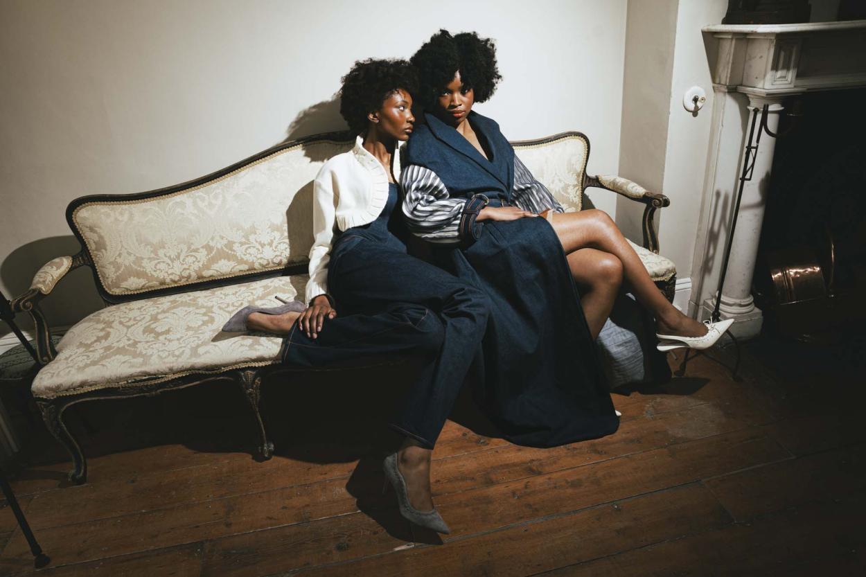 Two models posed sitting on an antique couch, in long denim coat with stripe silk chiffon embellishment on báinín sleeves, and denim trousers, corset and báinín jacket.