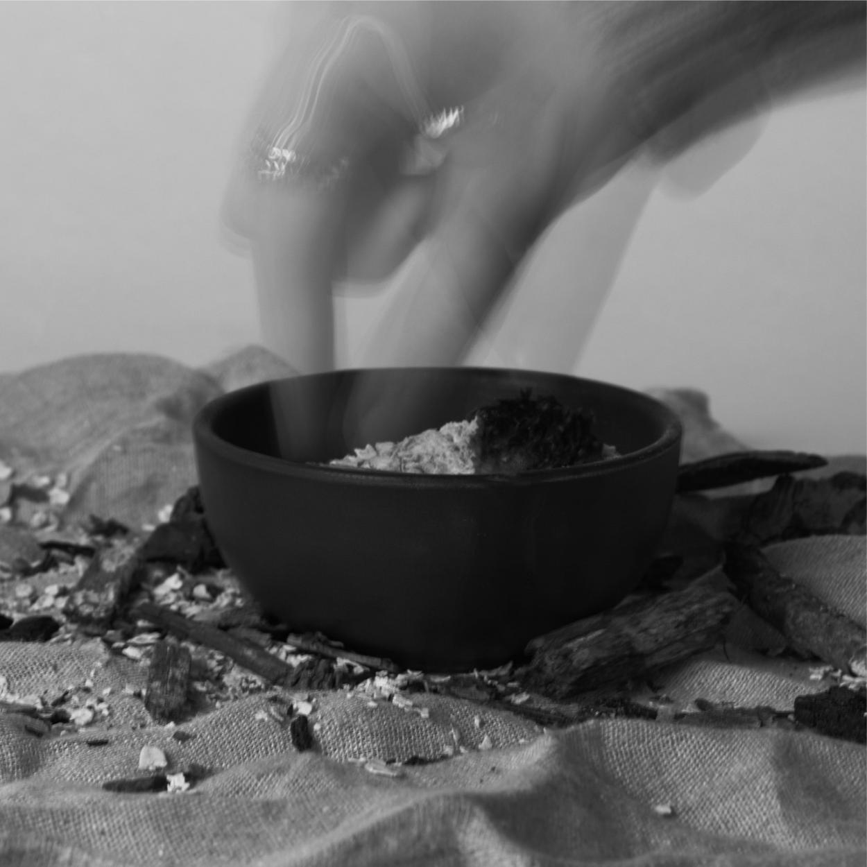 A black-and-white, long-exposure photograph capturing the blurred motion of a hand reaching into a dark bowl filled with oats on a rustic linen cloth.