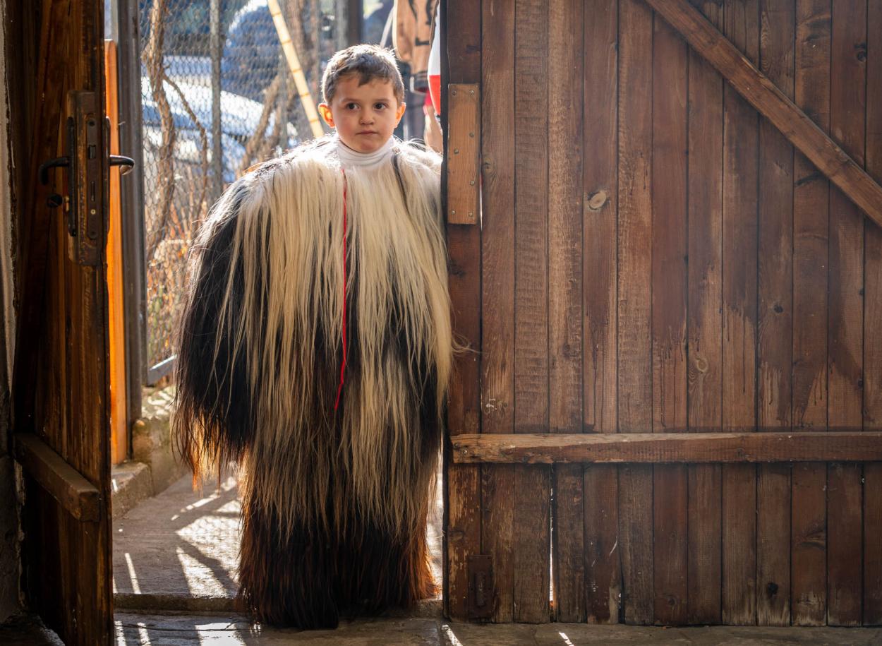 A child wearing a long, shaggy, two‑tone fur costume stands in a doorway with sunlight behind them.