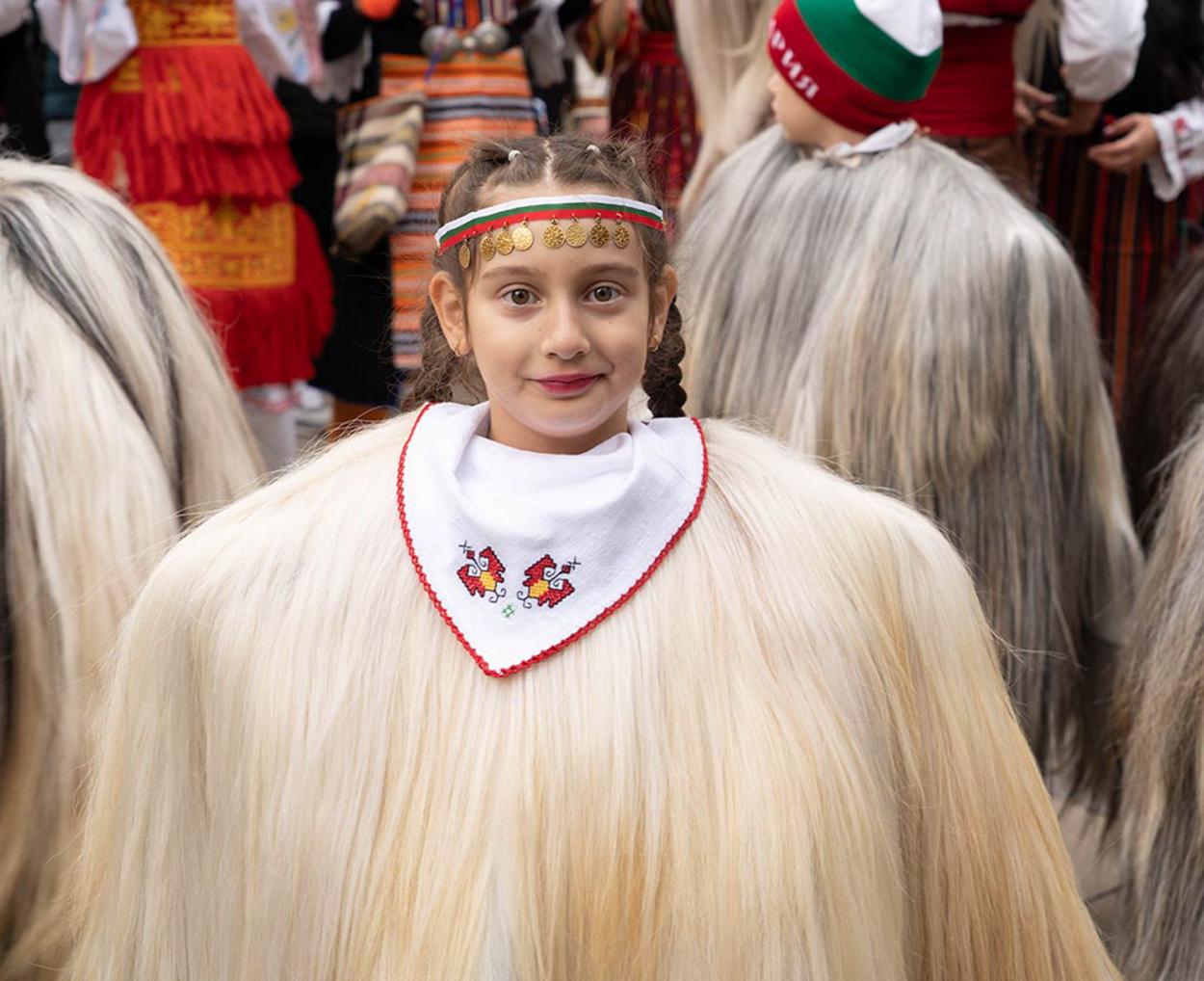 A girl in a white, long‑haired traditional costume stands smiling among others in festive attire.