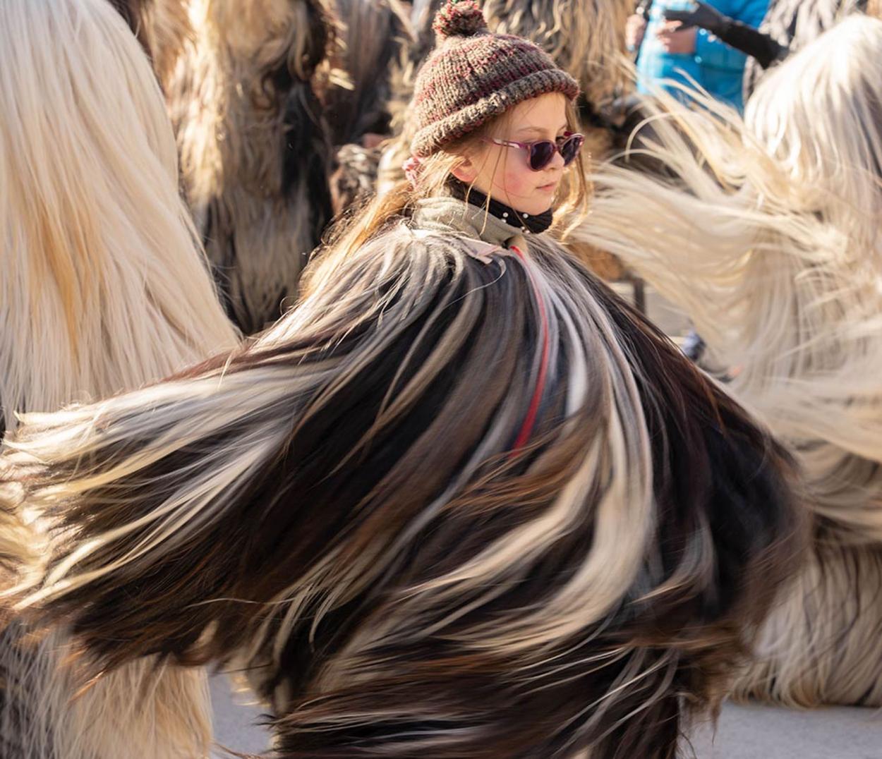 A child in a swirling, multi‑toned fur costume moves through a crowd, wearing a knit hat and sunglasses.