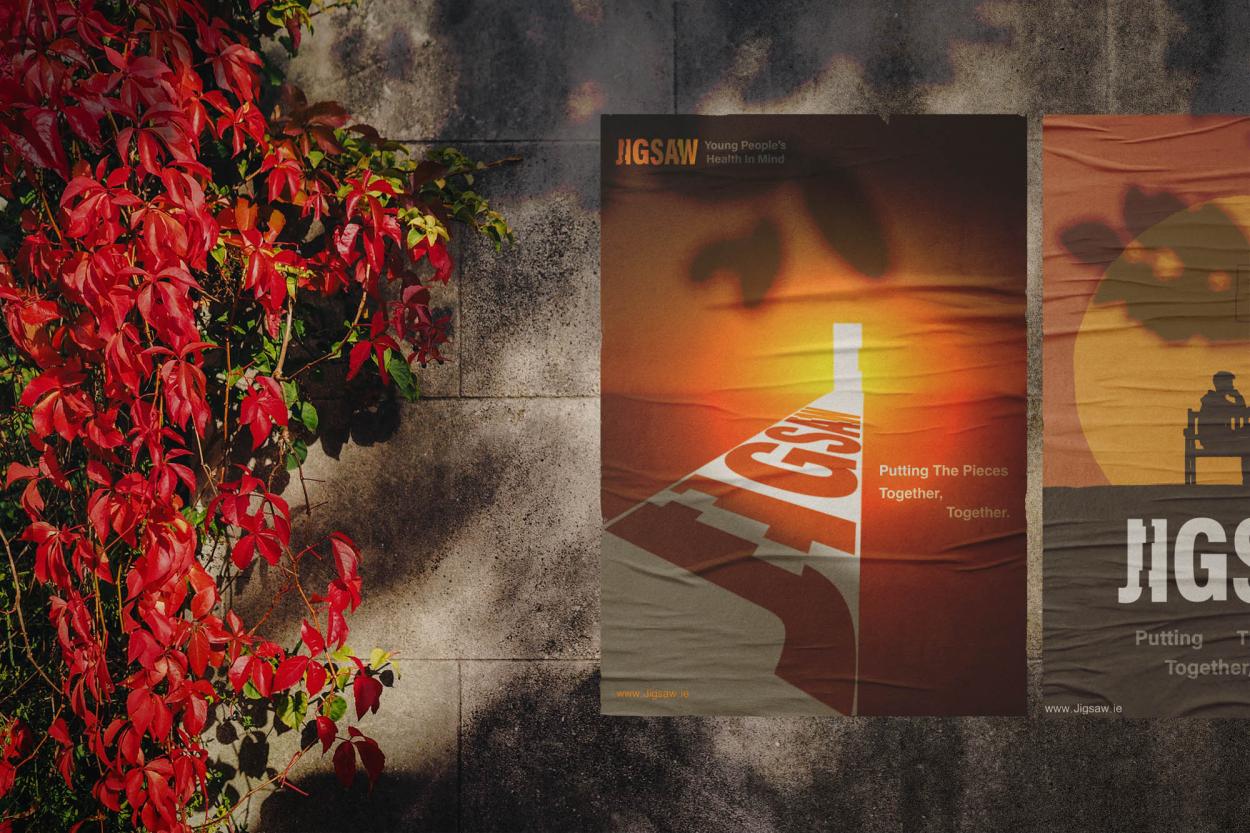 1-	Two Jigsaw posters are displayed on a concrete wall partially covered with vibrant red flowers. The main poster features a beam of light leaving a bright doorway. The Jigsaw logo and the tagline “Young People’s Health In Mind” appear at the top, alongside the message “Putting the Pieces Together, Together.” With a second poster beside it