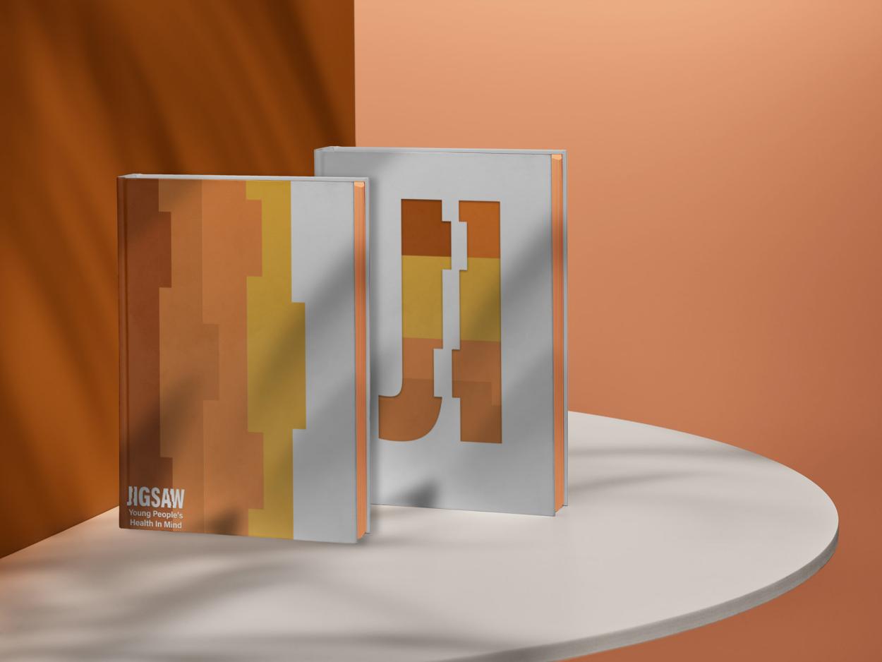 6-	2 journals are displayed on a white table in an orange lit room, the front and back cover of the journal are displayed showing interlocking puzzle piexes in a graident of orange and yellows with a shortened version of the Jigsaw logo.