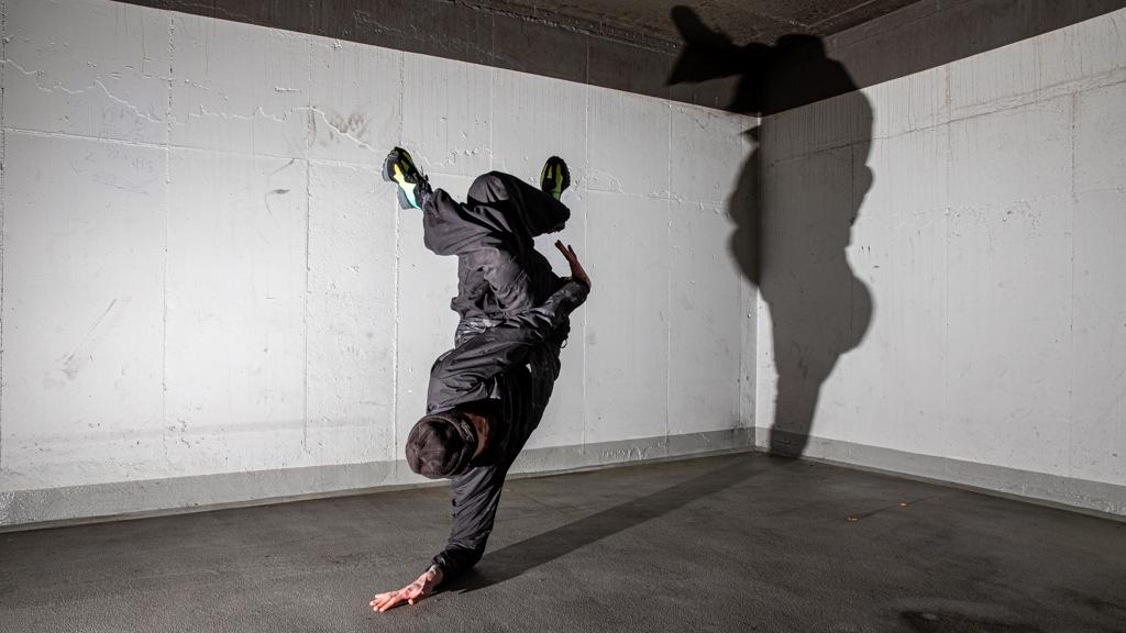 A person in black holds a one-handed freeze, legs extended toward the wall, with a bold shadow cast in a concrete corner.