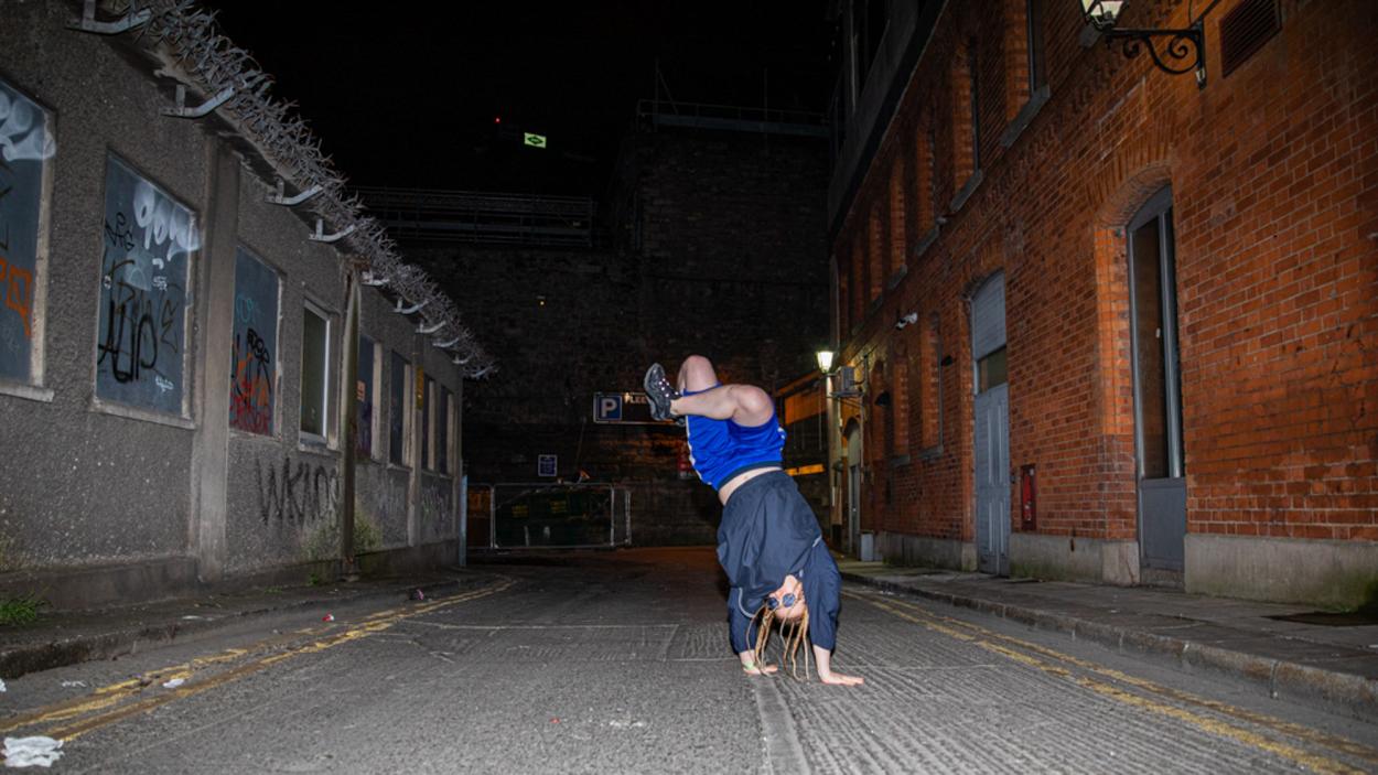 A person in a blue tank top performs a handstand in a graffiti-lined alley at night, brick buildings flanking both sides.