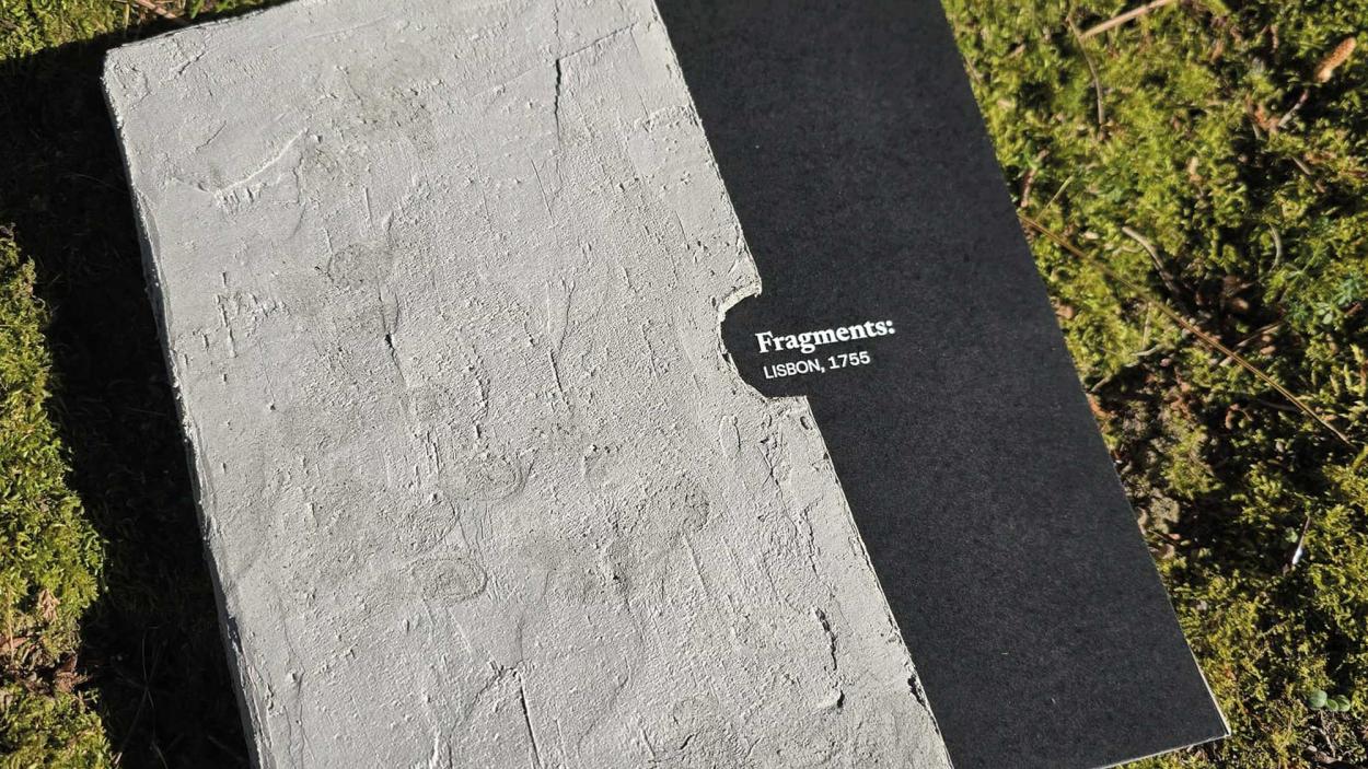 A black book titled "Fragments: Lisbon, 1755" sits inside a textured, concrete-like case on vibrant green moss