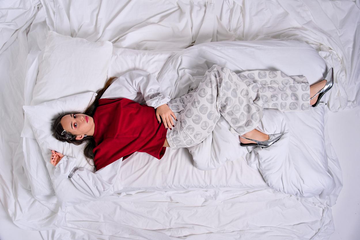 Top view of a model laying in a white bed, wearing a red and white outfit with printed jeans