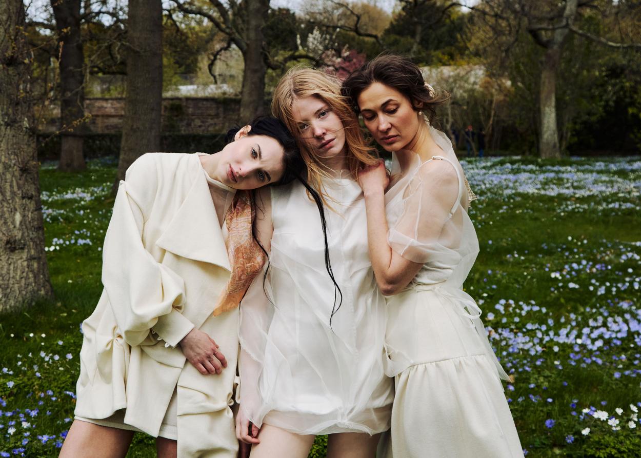 Three models standing together in a flower field dressed in wool and sheer silk outfits.