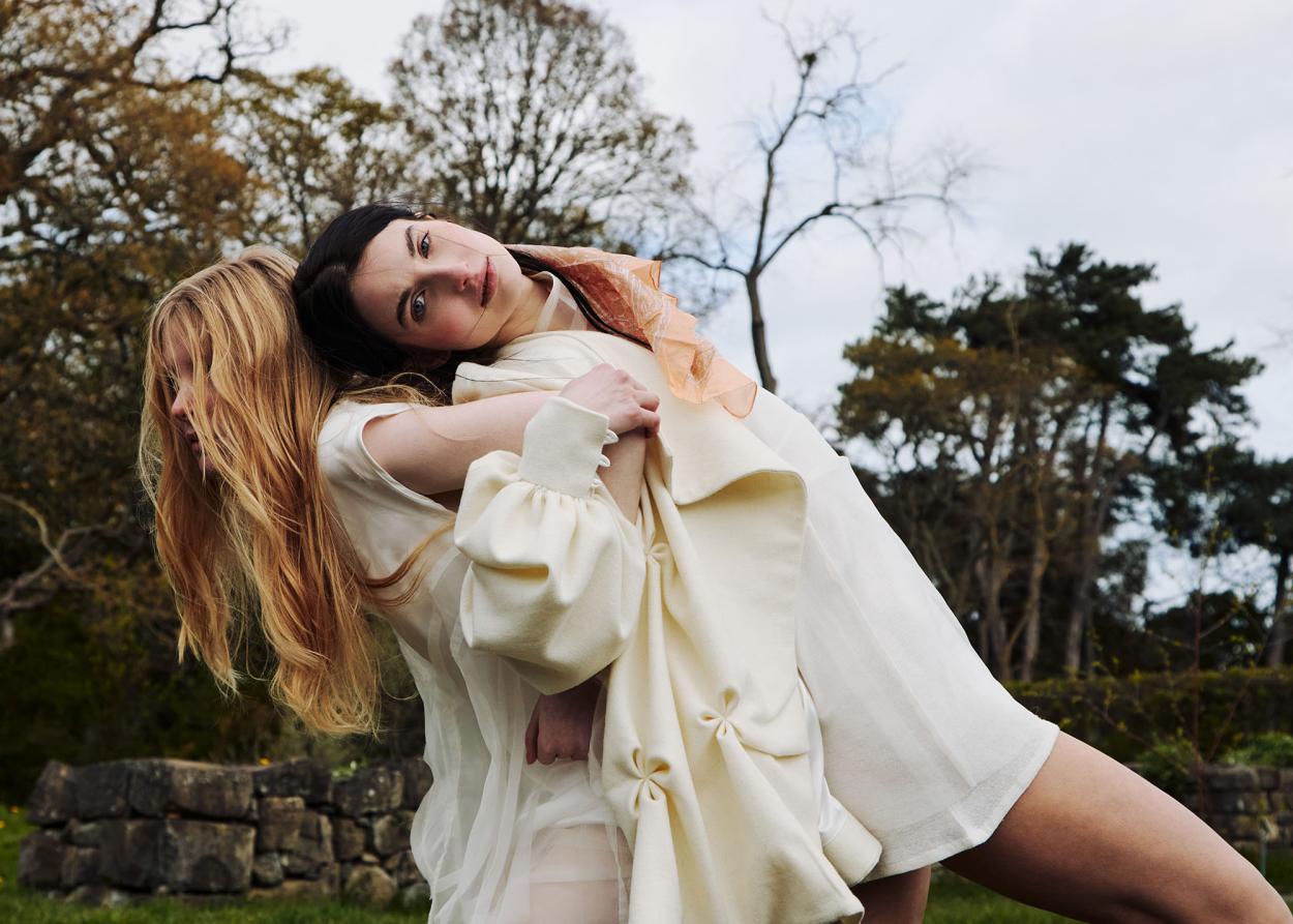 Two models back-to-back with one leaning on the other and trees in the background.