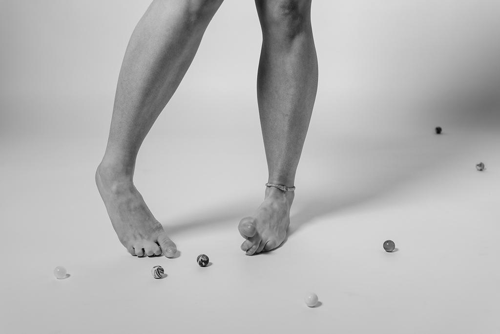 A black-and-white close-up of bare feet balancing marbles on the floor, with scattered marbles surrounding them.