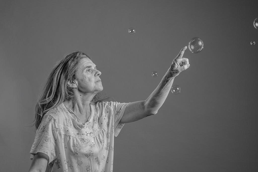  An older woman reaches towards floating soap bubbles in a black-and-white portrait against a plain background.
